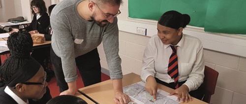 Blueprint for All photo (a man with glasses leans over a desk and helps two schoolgirls with a project)